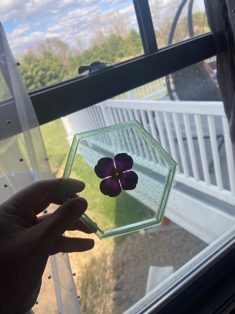 Puede incluir: Un posavasos hexagonal de resina verde y blanca con una flor morada prensada en el centro.