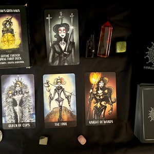 Puede incluir: Una baraja de tarot de drag de fantas&iacute;a g&oacute;tica con 75 cartas y una gu&iacute;a digital. La caja es negra con un dise&ntilde;o de telara&ntilde;a blanca y el texto "Hannah's Goth Haus". Las cartas presentan ilustraciones de drag queens con trajes de fantas&iacute;a g&oacute;tica.