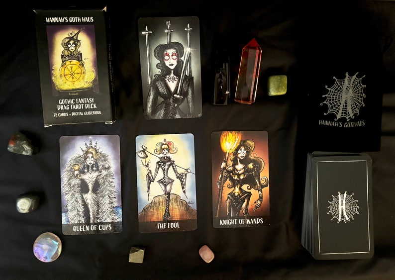 Puede incluir: Una baraja de tarot de drag de fantas&iacute;a g&oacute;tica con 75 cartas y una gu&iacute;a digital. La caja es negra con un dise&ntilde;o de telara&ntilde;a blanca y el texto "Hannah's Goth Haus". Las cartas presentan ilustraciones de drag queens con trajes de fantas&iacute;a g&oacute;tica.