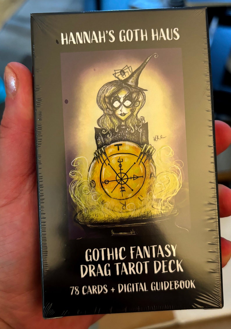 Puede incluir: Una caja negra con una ilustraci&oacute;n colorida de una bruja sosteniendo una carta de tarot. El texto en la caja dice "Hannah's Goth Haus Gothic Fantasy Drag Tarot Deck 78 Cards + Digital Guidebook".