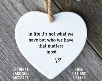 May include: A white heart-shaped ornament with a jute string for hanging. The ornament has the text "in life it's not what we have but who we have that matters most" and a small heart symbol. The background is a wooden surface.