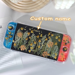 May include: A clear protective case for a gaming device, featuring a black background with a floral design of dried yellow and orange flowers. The name "Daisy" is printed on the case. The device's controls are blue and red.
