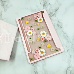 Puede incluir: Una funda rosa para tableta con una parte delantera transparente adornada con flores prensadas en tonos rosa, amarillo y blanco. La funda incluye un soporte para lápiz y una tapa plegable. Una caja rosa a juego es visible en el fondo.