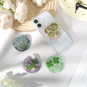 May include: Heart-shaped phone grips with dried flowers and greenery encased in clear resin. One grip is attached to a phone in a clear case. Other grips feature a four-leaf clover, fern, and other botanical elements.