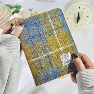 May include: A rectangular, patterned notebook cover in shades of blue, gold, and brown. The cover features a plaid design with a white accent. A small label is attached to the cover. The cover is held by a person.