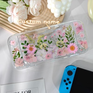 May include: Clear protective case for a gaming console, adorned with pressed pink and white flowers. The case has a rectangular shape and is designed to fit the console. The text "Custom name" is visible on the case.