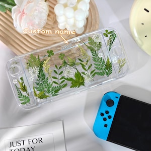 Może przedstawiać: Przezroczyste etui na Switch z prasowanymi zielonymi liśćmi i białymi kwiatami. Etui ma niestandardową etykietę z nazwą. Widoczny jest niebiesko-czarny kontroler Switch. Akcesorium dekoracyjne do konsoli do gier.