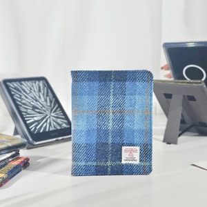 May include: A blue plaid Kindle cover with a rectangular shape, featuring a woven fabric texture. The cover has a small label on the front. Other Kindle covers and e-readers are visible in the background.