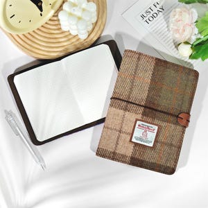 Puede incluir: Un cuaderno abierto con papel cuadriculado junto a un cuaderno cerrado con una cubierta a cuadros marrón y verde. El cuaderno cerrado tiene una correa de cuero y una etiqueta que dice "Harris Tweed". También se ve un bolígrafo transparente.