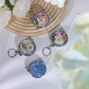 May include: Four small, round, silver-toned compact mirrors with floral designs. One mirror features blue flowers and rhinestones. The other three mirrors have pressed flowers in various colors. Some mirrors have keychains attached.