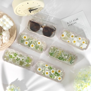 Può includere: Custodie rettangolari trasparenti piene di fiori secchi e verde. Una custodia contiene occhiali da sole. Le custodie presentano margherite, trifogli e altre piante. È visibile un foglio con il testo "JUST FOR TODAY".