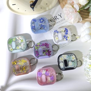 Może przedstawiać: Kolekcja przezroczystych, kwadratowych etui na AirPods, każde wypełnione suszonymi kwiatami w różnych kolorach. Etui posiadają srebrny brelok. Kolory obejmują niebieski, zielony, fioletowy, żółty, różowy i czarny.