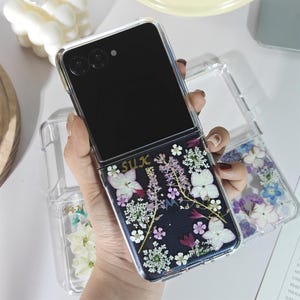 May include: A clear phone case with a black phone inside. The case is decorated with dried flowers in white, purple, and pink against a dark background. The word "SULX" is printed in gold.
