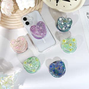 Puede incluir: Una colección de soportes para teléfono en forma de corazón en varios colores, incluyendo rosa, morado, verde y azul. Cada soporte presenta diseños florales o con purpurina y algunos tienen texto. Un soporte está adherido a una funda transparente.