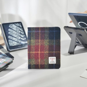 May include: A Harris Tweed tablet case in a plaid design, featuring red, blue, and yellow colours. The case is shown with an e-reader, a tablet on a stand, a clock, and flowers. The case is approximately 20 cm in height.