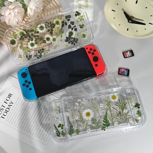 May include: A Nintendo Switch console with a black screen and red and blue controllers, inside a clear case decorated with pressed flowers. Another case with a similar design is above. Two game cartridges are visible.