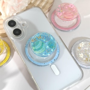 May include: A clear phone case with a blue and green glitter phone grip attached. The grip has a celestial design with gold accents. Other phone grips with pink, yellow, and white designs are also visible.