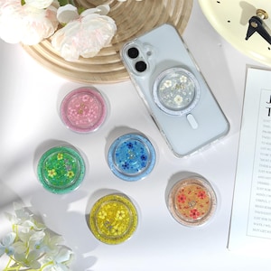 May include: A collection of colorful phone grips with floral designs. The grips are round and transparent, filled with dried flowers in pink, green, blue, yellow, and orange. One grip is attached to a clear phone case. The background includes a clock and a piece of paper.