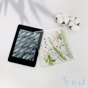 Puede incluir: Una bandeja cuadrada de resina transparente con flores y vegetación prensadas en su interior. La bandeja está sobre una superficie blanca junto a un lector electrónico negro.