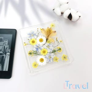 Op de afbeelding: Een vierkant, transparant frame met geperste bloemen erin. De bloemen zijn geel, wit en blauw. Het frame staat op een wit oppervlak.