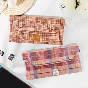 Op de afbeelding: Twee rechthoekige stoffen etuis met klepsluitingen. De bovenste etui is een roodachtig-roze tweed met goudkleurige knoopsluitingen. De onderste etui heeft een ruitpatroon in roze, rood en blauw met goudkleurige knoopsluitingen.