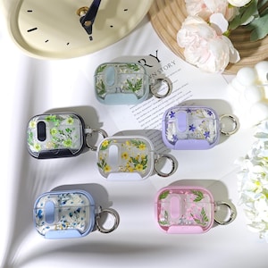Può includere: Cinque custodie rettangolari per auricolari con coperchi trasparenti contenenti fiori e vegetazione pressati. Ogni custodia presenta una base in tinta unita nera, azzurra, bianca, lavanda o rosa e include un anello in metallo color argento.