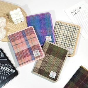 May include: Four rectangular notebook covers featuring various plaid patterns in pink, blue, beige, and green tones. Each cover has a small white rectangular label with text and a stitched border. The covers are arranged on a white surface.