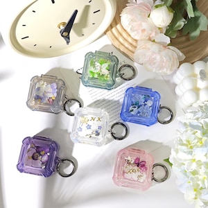 May include: A collection of clear, square-shaped AirPod cases with floral designs and keychains. The cases are in various colors, including purple, blue, green, and pink. Some cases have the letters "NX" or "CO" on them. The background includes a clock and flowers.