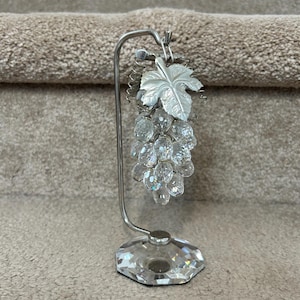 May include: A decorative crystal grape cluster sculpture with a silver-colored metal stand. The grapes are made of clear, faceted crystals, and the stand has a leaf detail. The base is a faceted crystal. The sculpture is approximately 6 inches tall.