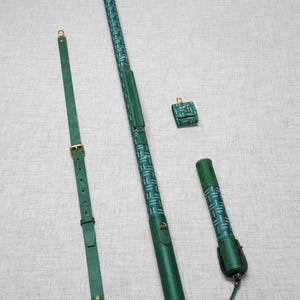 Puede incluir: Un conjunto de accesorios de cuero verde, que incluye un estuche largo, una pequeña bolsa, un estuche más corto y una correa para el hombro. Los estuches largos y cortos tienen un patrón geométrico en un tono verde más claro. La correa tiene una hebilla y herrajes dorados.