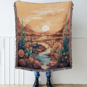 May include: A woven tapestry throw blanket featuring a desert landscape with cacti, a stream, and a sunset. The colors are muted and earthy, with shades of brown, green, and orange.