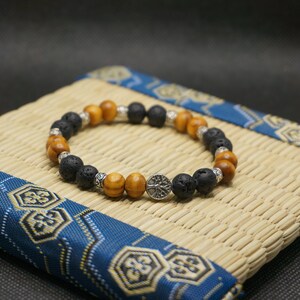 Peut inclure: Un bracelet composé de perles en bois brun et de perles en lave noire. Le bracelet est orné d'un charme en argent avec un motif d'arbre.
