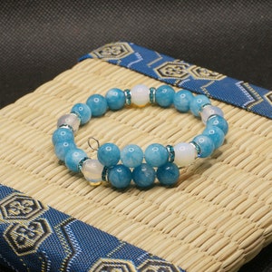 Op de afbeelding: Een blauwe en witte kralen armband met een geweven achtergrond. De armband is gemaakt van ronde, lichtblauwe kralen met kleinere, witte kralen die ertussen zijn gestoken.