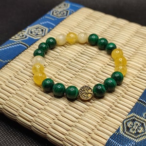 Peut inclure: Un bracelet en perles vertes, jaunes et blanches avec un charme en or en forme d'arbre de vie. Le bracelet est sur un tapis tissé.