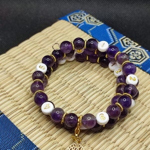 Peut inclure: Un bracelet double brin en améthyste avec des accents dorés et des perles blanches avec des chiffres dorés. Le bracelet a un charme d'arbre en or.