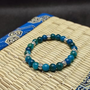 Puede incluir: Un brazalete de piedras preciosas azules y verdes con detalles plateados. El brazalete está hecho de cuentas redondas y está enhebrado en un cordón elástico.