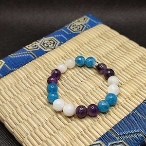 Puede incluir: Un brazalete hecho con cuentas blancas, azules y moradas. El brazalete tiene un dije de plata.