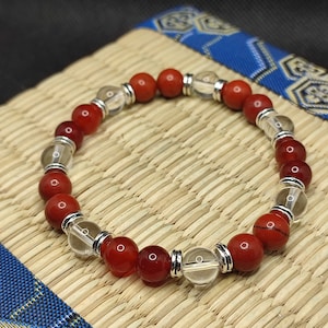 Puede incluir: Un brazalete de cuentas de cornalina roja y cuarzo transparente con detalles plateados. El brazalete está sobre una estera de paja tejida.