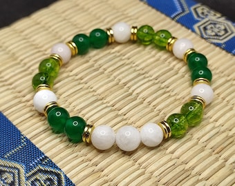 May include: A green, white, and gold beaded bracelet with gold spacers. The bracelet is made with round beads and is lying on a woven mat.