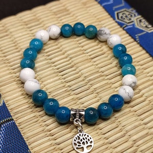Puede incluir: Un brazalete de cuentas azules y blancas con un dije de árbol de la vida plateado. El brazalete está hecho con cuentas redondas y tiene una banda elástica.
