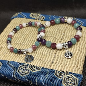 Op de afbeelding: Twee armbanden met kralen van een verscheidenheid aan kleurrijke edelstenen, waaronder wit, paars, groen en rood. Elke armband heeft een zilveren bedel, een met een levensboomontwerp en de andere met het woord "LOVE". De armbanden worden gepresenteerd op een geweven mat.