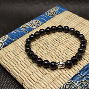 Peut inclure: Bracelet en perles noires avec un charme Bouddha argenté. Le bracelet est sur un tapis tissé avec une bordure à motifs bleus et blancs.