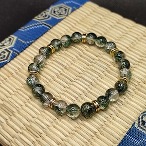 Könnte beinhalten: Ein grünes und klares Kristall-Armband mit goldenen Akzenten. Das Armband besteht aus runden Perlen und ist auf einer elastischen Schnur aufgefädelt.
