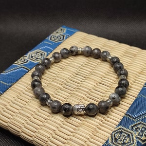 Peut inclure: Un bracelet en pierres grises avec un charme en argent en forme de Bouddha. Le bracelet est composé de perles rondes et a un design simple.