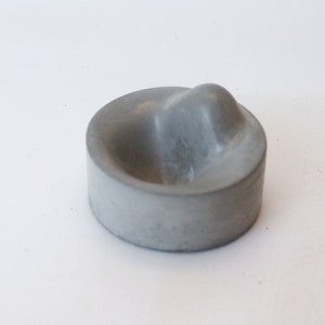 May include: A small, gray concrete bowl with a rounded, raised center. The bowl is smooth and has a slightly textured surface.