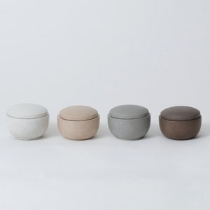 May include: Four round ceramic containers with lids in various shades of gray, beige, and brown. The containers are arranged in a row from left to right.