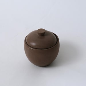 May include: A small, brown ceramic jar with a lid. The jar is round with a slightly textured surface and a small knob on top of the lid. The jar is set against a white background.