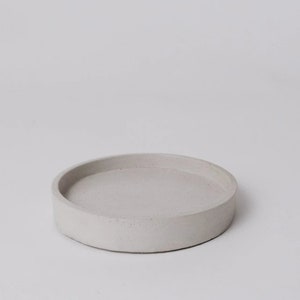 May include: A round, white concrete tray with a slightly raised edge. The tray is smooth and has a subtle texture.