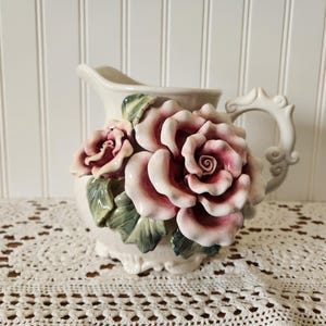 May include: Cream-colored ceramic pitcher with a sculpted rose design. The roses are a mix of pink and white, with green leaves. The pitcher has a decorative handle and a scalloped base, perfect for floral arrangements.