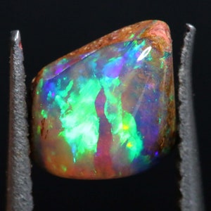 May include: A rough, natural opal gemstone with a vibrant green, blue, and purple iridescent sheen. The opal is a teardrop shape and is held in place by metal tweezers.